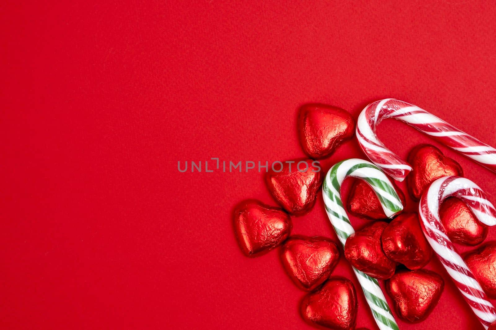 red festive Christmas background with candy in the shape of hearts wrapped in shiny foil and candy in the shape of a caramel cane