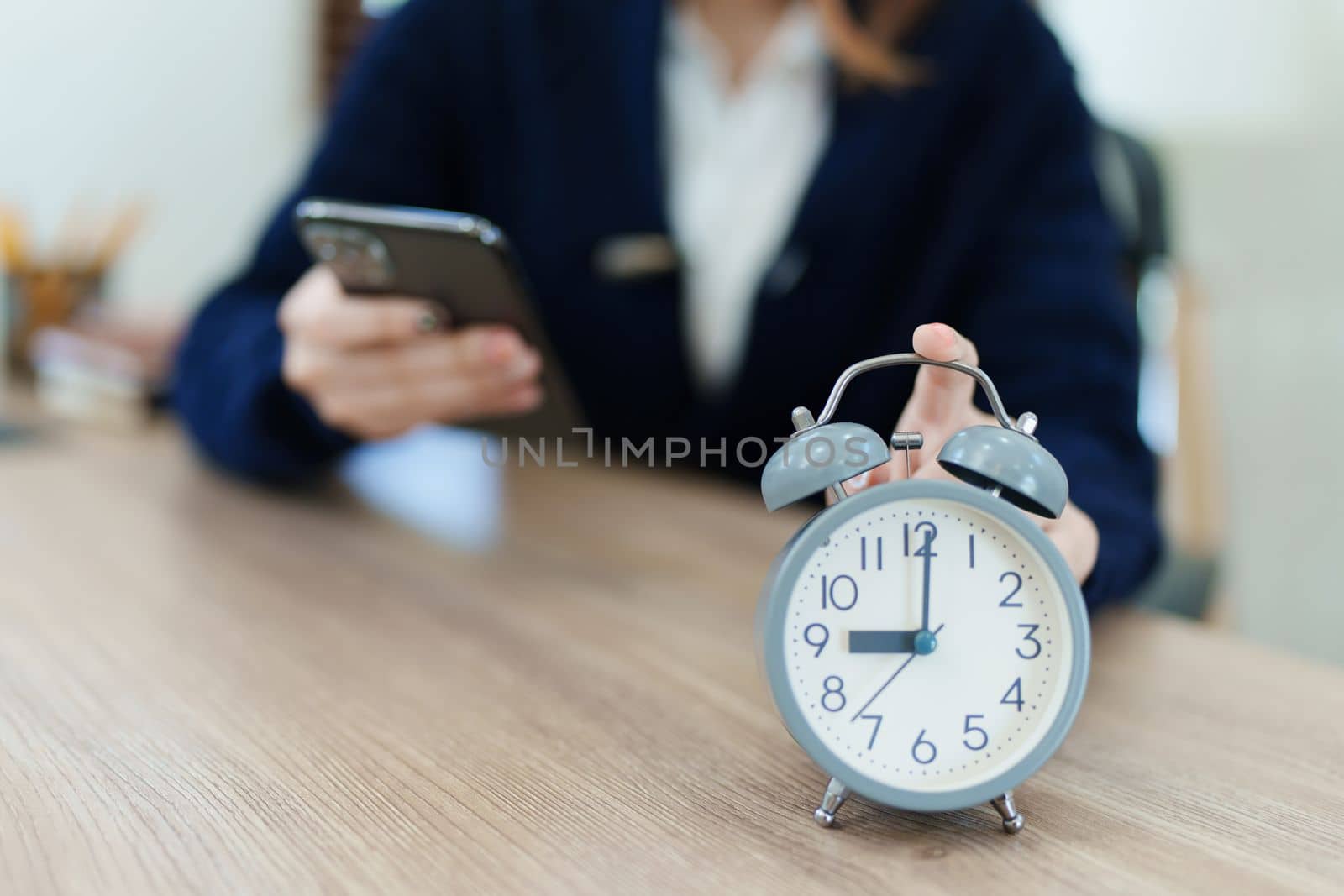 Alarm clock on the desk. Asian Businesswoman working in modern office building or home at night using smart phone at office by Manastrong