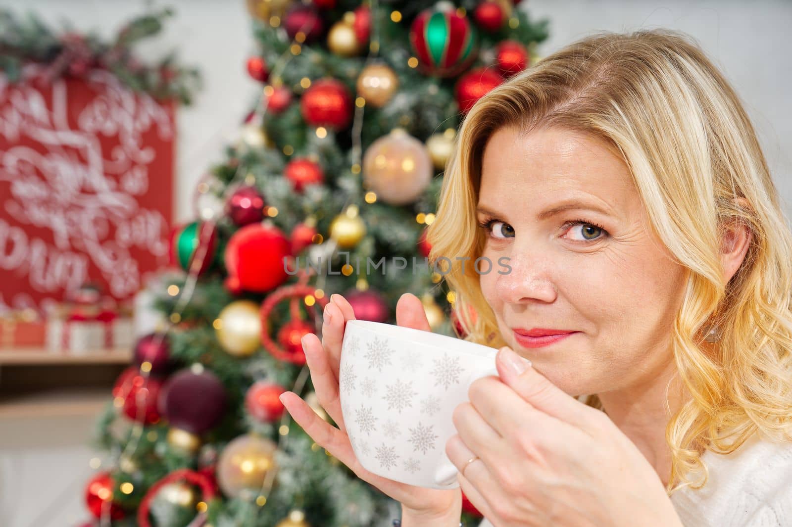 Portrait of woman 40s in white sweater holding a cup of warm coffee or tea on background of a Christmas tree. Cozy christmas atmosphere. warming non-alcoholic drink for Christmas. lifestyle