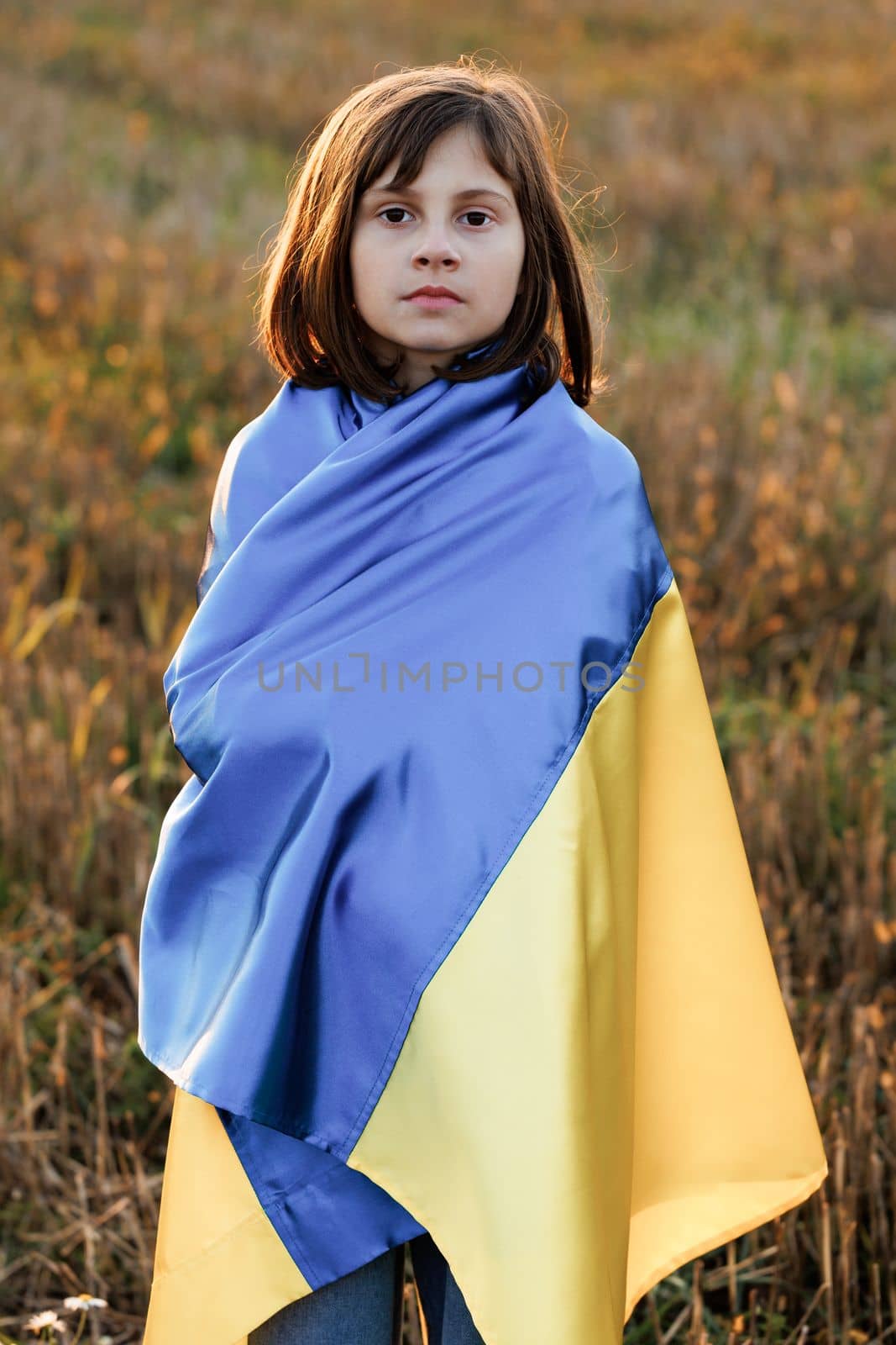 Portrait of Sad Ukrainian girl stands with a flag of Ukraine in the middle of field against a sunset sky. 8 years old girl holding the flag of Ukraine. War in Ukraine. Support for Ukraine.
