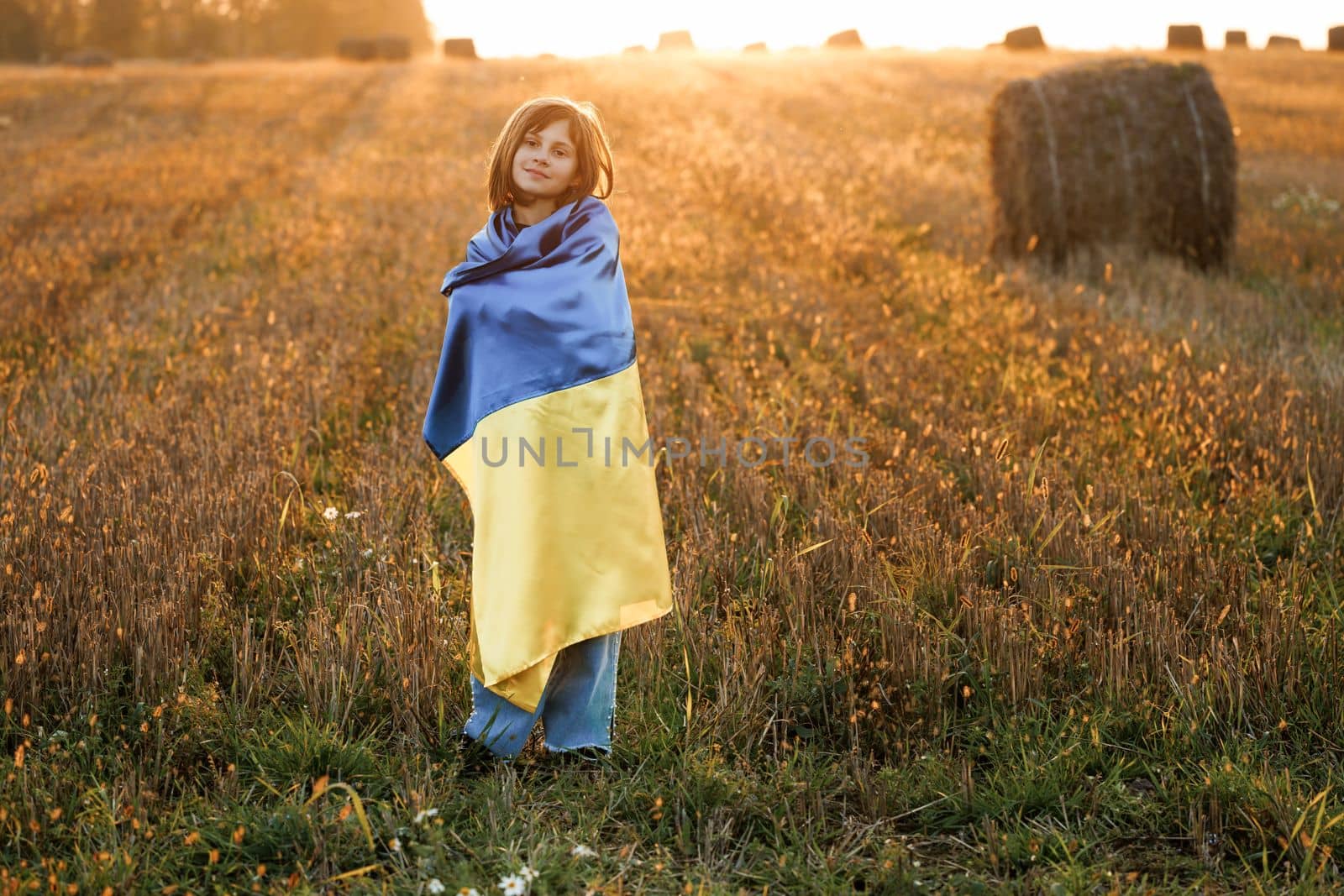 Portrait of Ukrainian girl stands with a flag of Ukraine in the middle of field against a sunset sky. Ukrainian girl with a blue-yellow flag against the sky. War in Ukraine. Ukrainian Independence Day