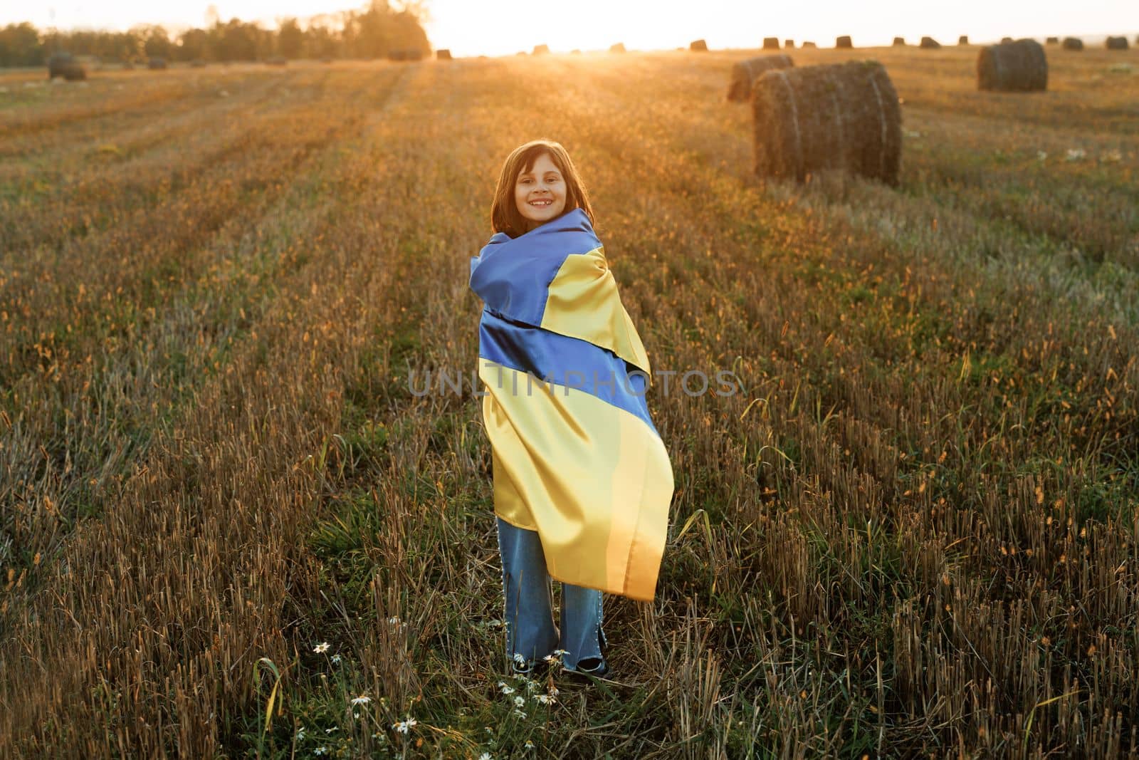 Ukrainian girl with a blue-yellow flag against the sky. War in Ukraine. Ukrainian Independence Day. Portrait of Ukrainian girl stands with a flag of Ukraine in the middle of field against a sunset sky