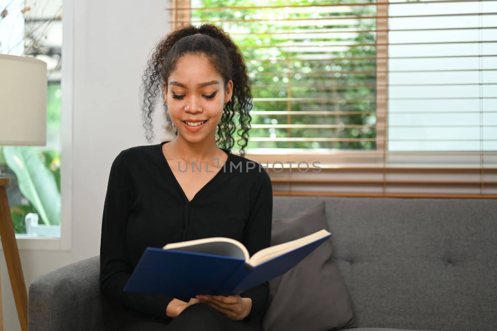 Young African American woman in casual clothes resting on couch and reading favorite literature by prathanchorruangsak