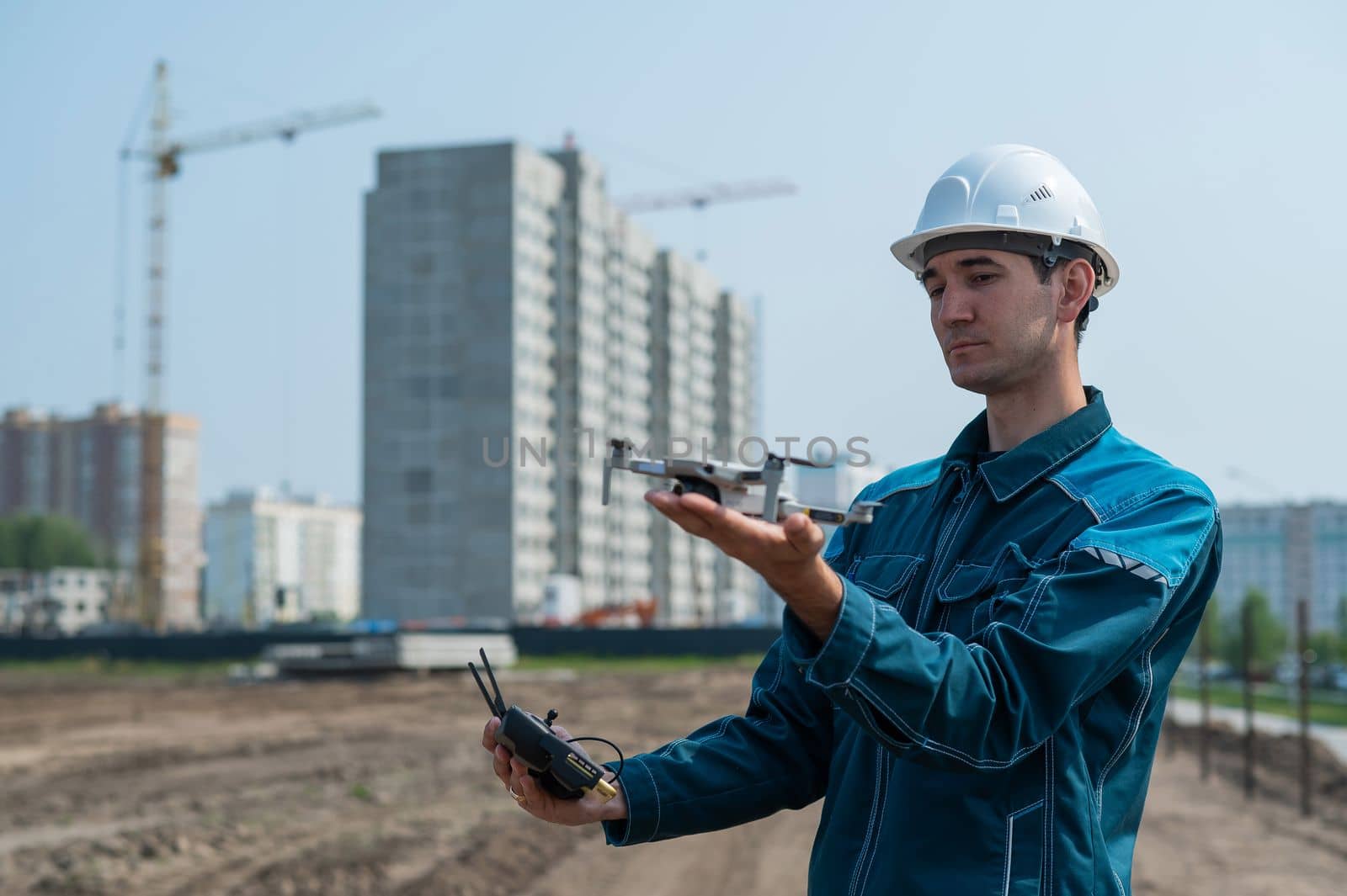 A man in a helmet and overalls controls a drone at a construction site. The builder carries out technical oversight