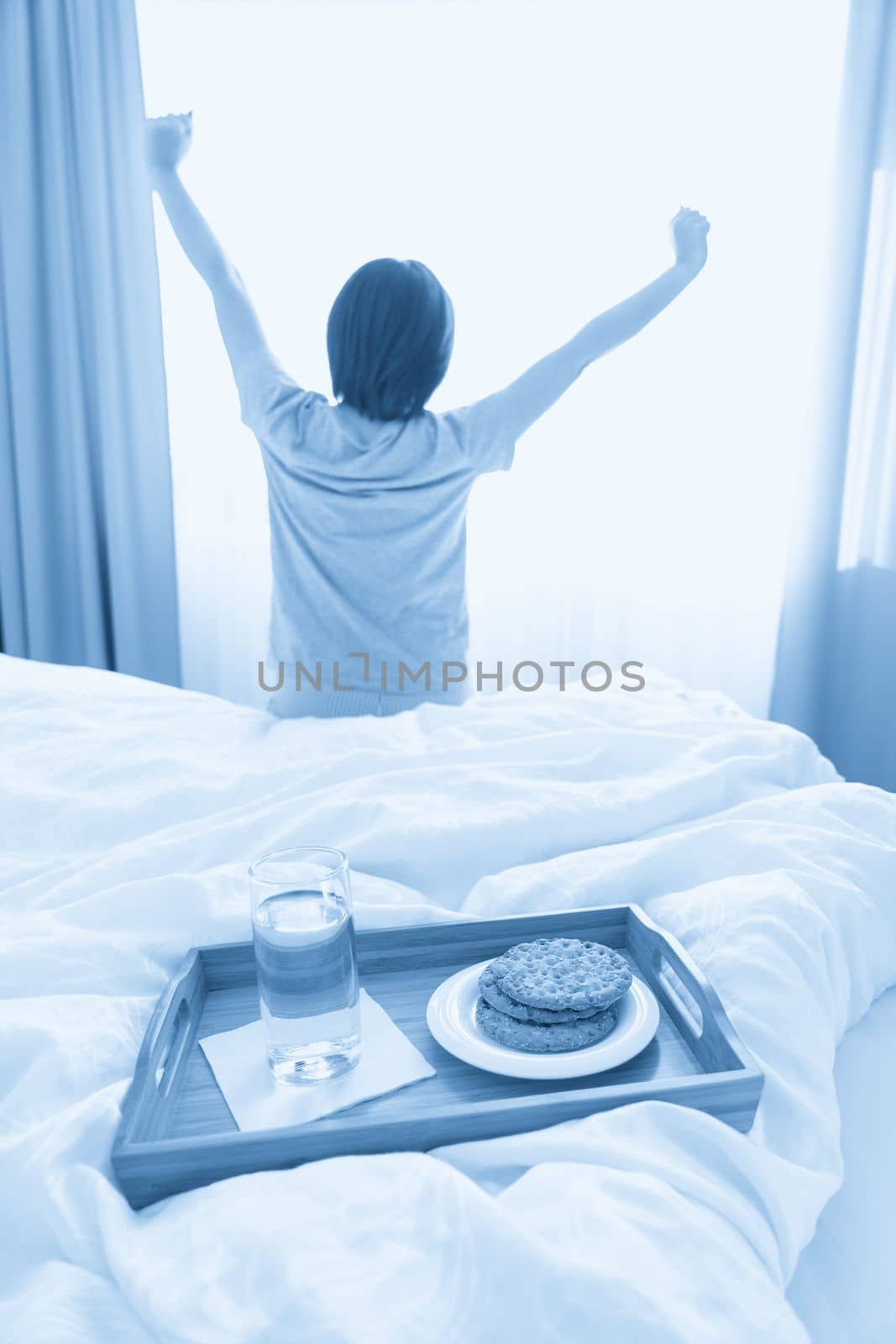 Tray with water and crackers breakfast on a bed with woman stretching on background by Mariakray