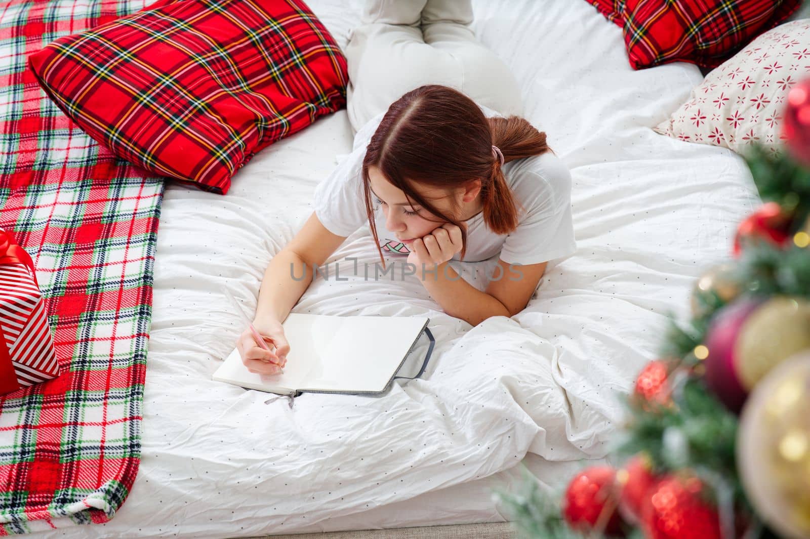happy teenage girl with pen and notebook making wish list or to do list for new year in bed over christmas tree. teenage girl dreaming in christmas time. xmas holidays concept