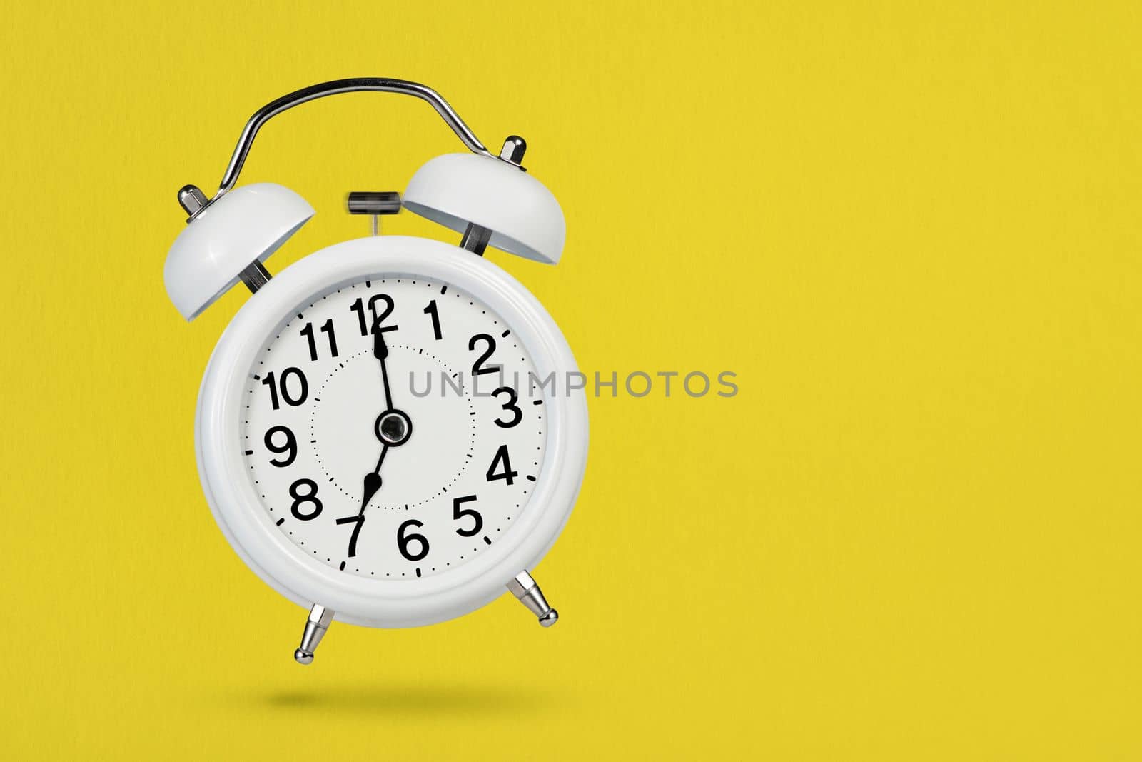 A white vintage alarm clock shows 7 o'clock, casting a shadow on a yellow background. The concept of morning and early awakening. Time to act or play sports. Place with copy space