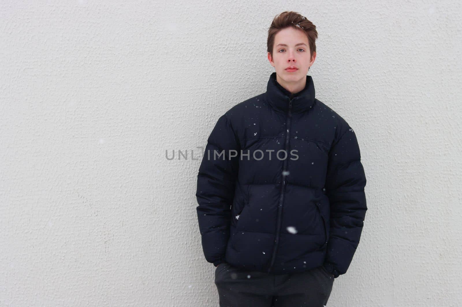 Seriously handsome teenage boy looking at the camera and holding his hands in his pockets against a backdrop of falling snowflakes. by gelog67