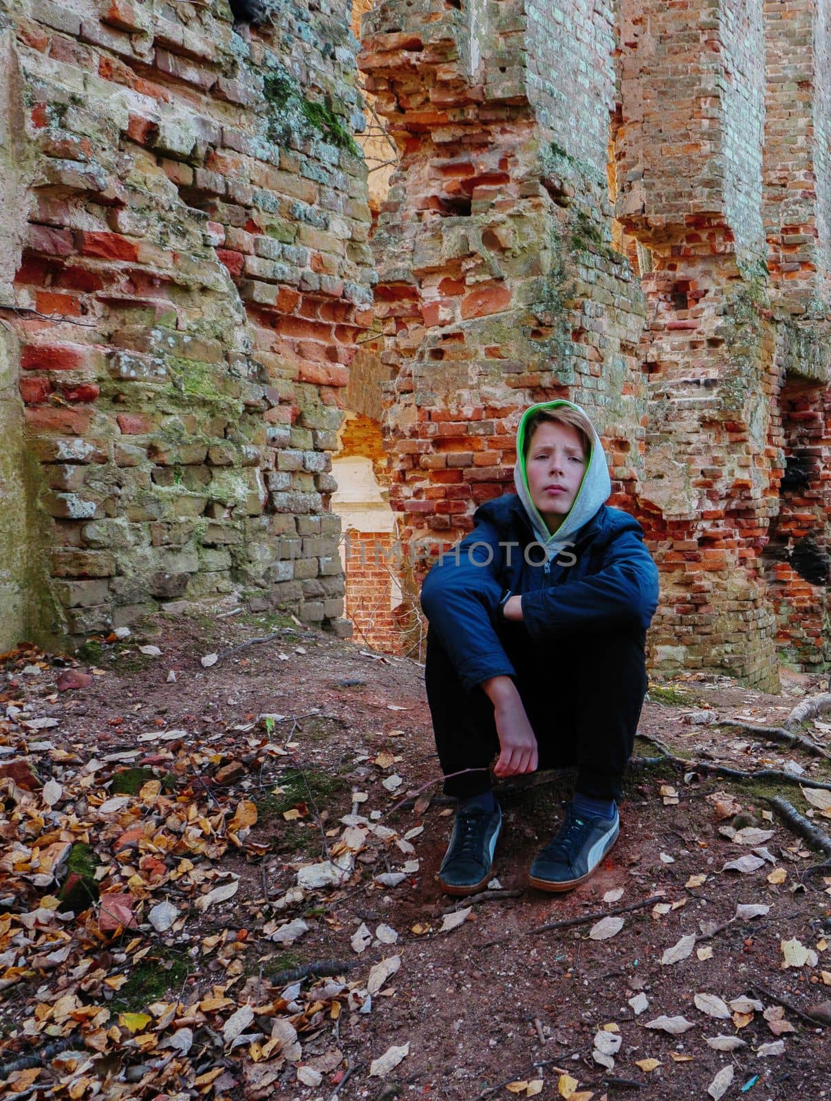A teenager in a blue jacket sits next to an old ruined brick wall in autumn. A sad boy of 2-15 years old sits on the ground near the destroyed red brick wall.