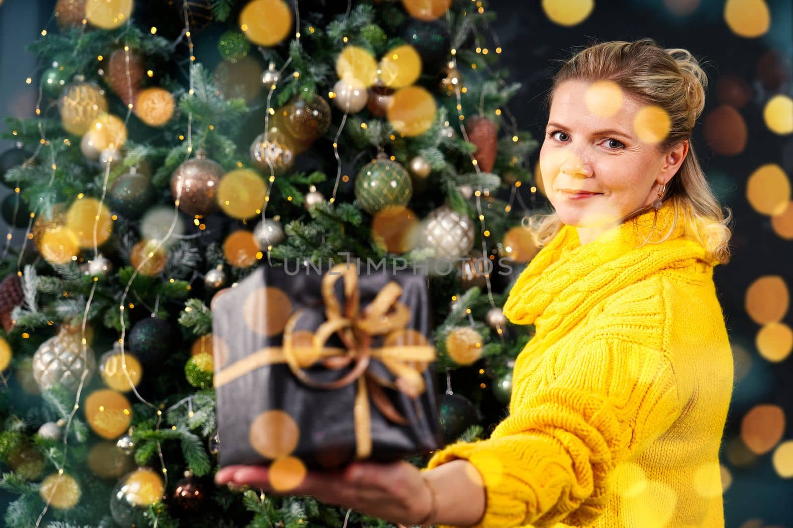 beautiful blonde woman holding a box with a christmas present in her hand. Decorated Christmas tree on background. Christmas lifestyle with copy space