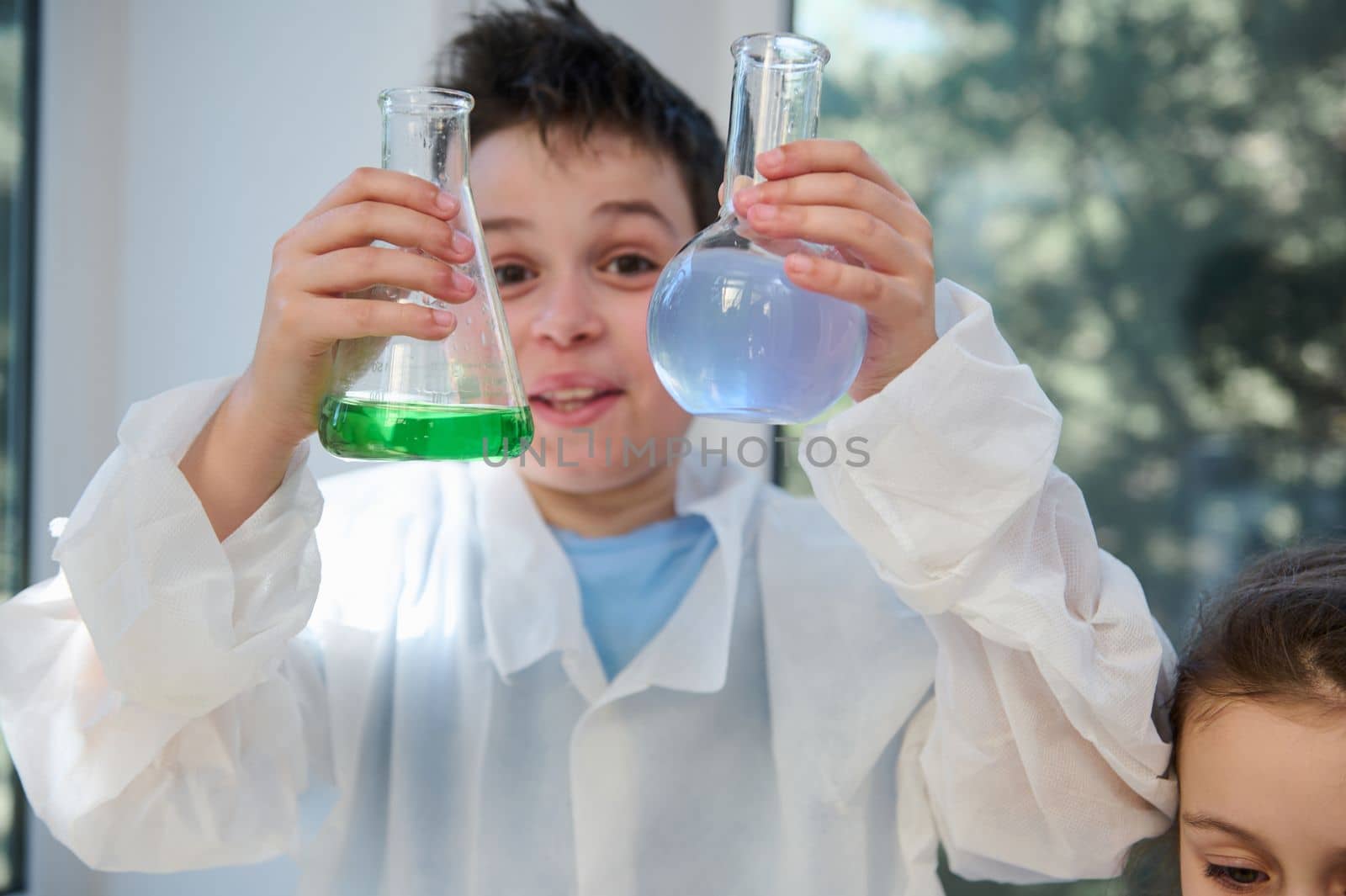 Details: selective focus on the hands of a schoolboy in white lab coat, holding lab flasks with chemical solutions, examining the liquid inside the lab glassware, in a school chemistry laboratory