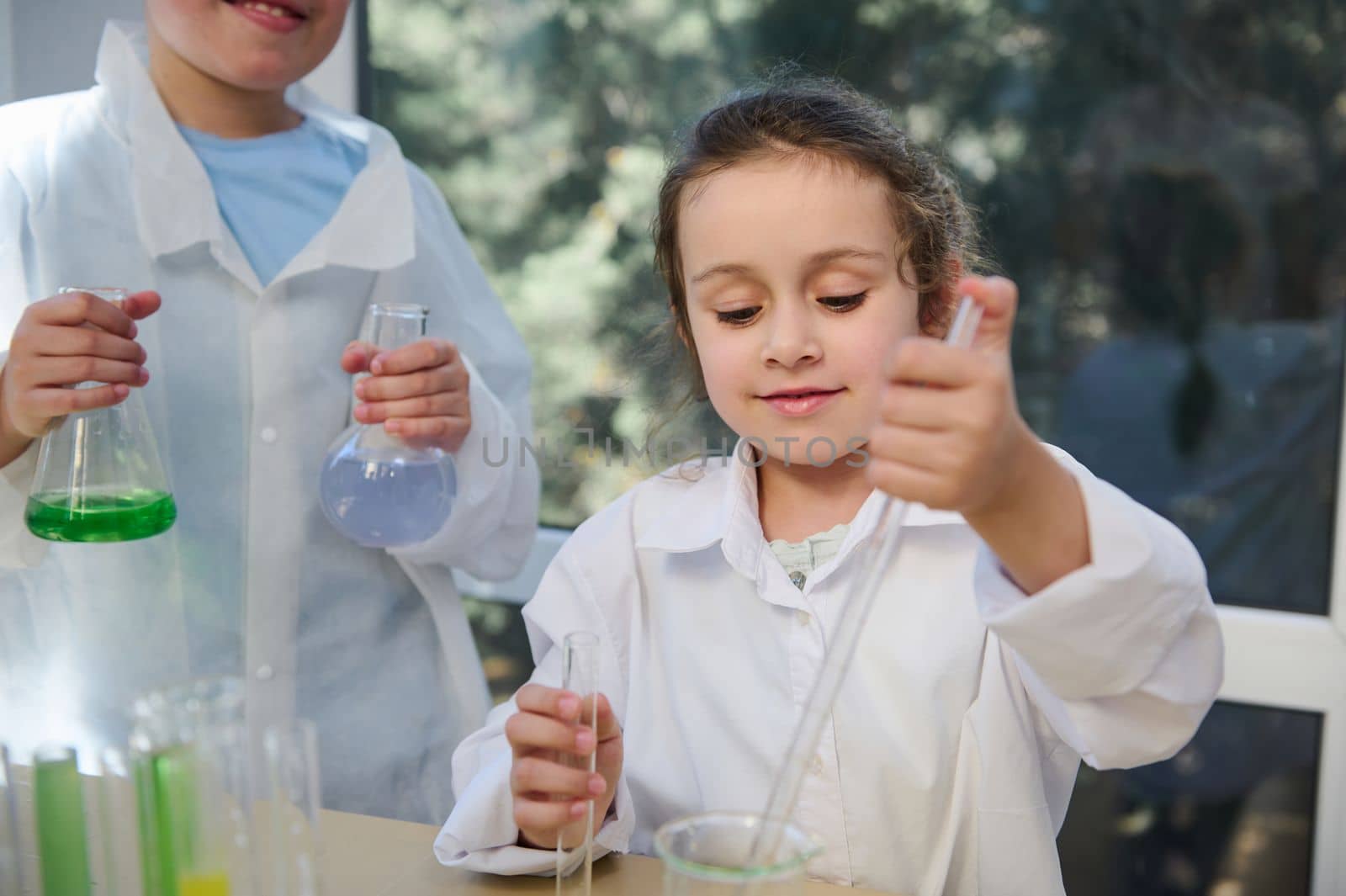 Caucasian 5-6 years old child, smart and beautiful smiling little school girl, enjoying chemistry class in school laboratory. Kids conducting chemical experiments. Learning new educational subject