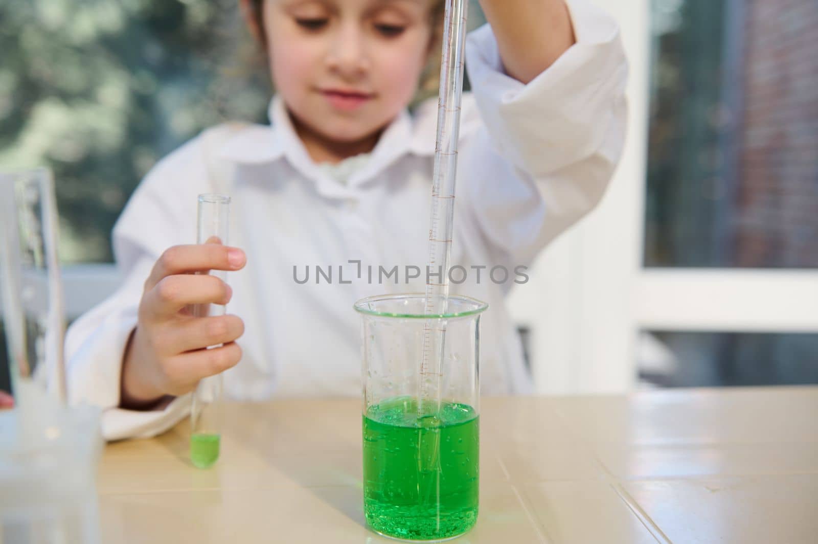 Selective focus on a glass lab beaker with green chemical liquid and graduated pipette in the hands of a blurred smart school girl, filling few reagent during chemistry class in the school laboratory