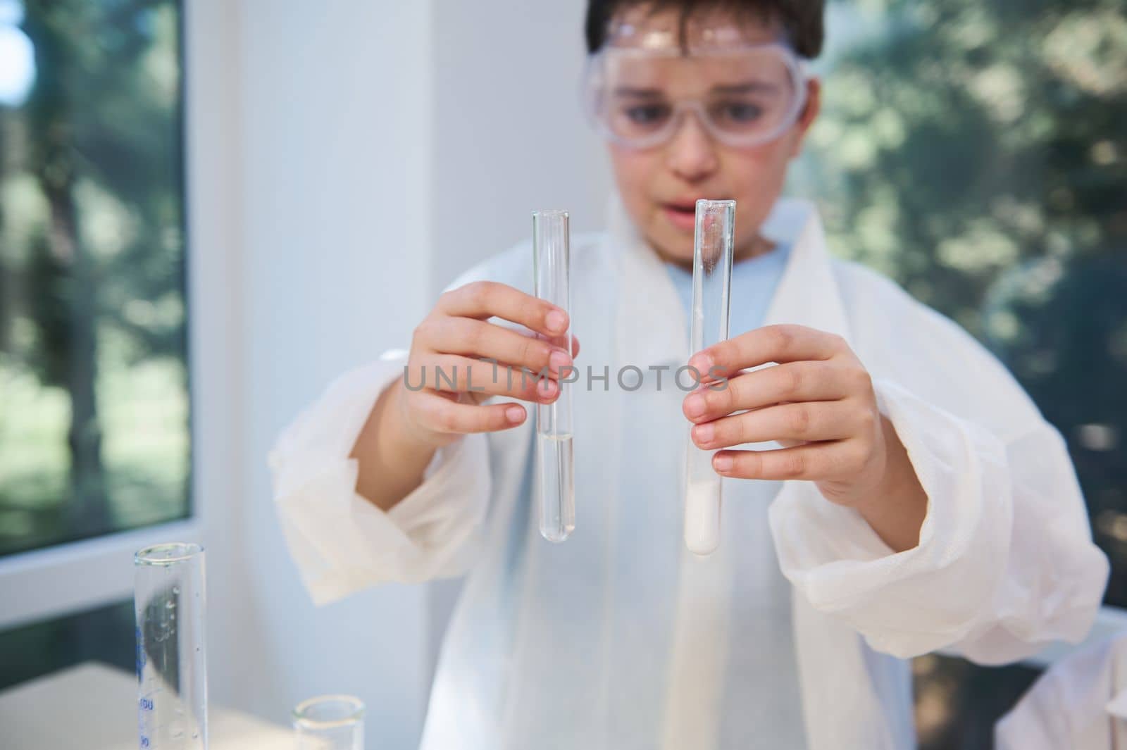 Selective focus on test tubes with chemicals and reagents, in the hands of a blurry smart teenage boy, wearing white lab coat and goggles, while a chemistry class in the school laboratory. Science