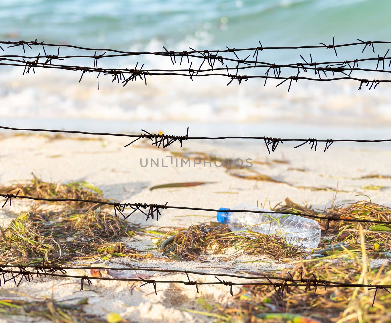 View through the barbed wire on a dirty beach filled with seaweed, garbage and empty plastic bottle on background