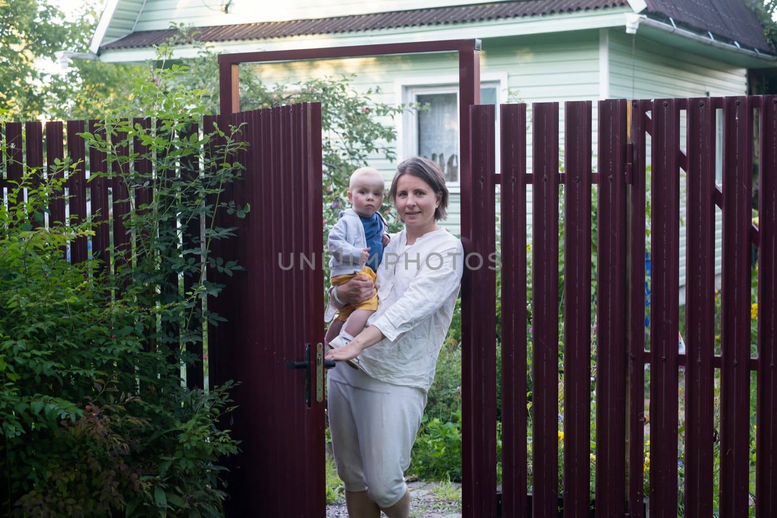 Curious woman with a baby opening the gate of fence and looking at camera welcoming friends