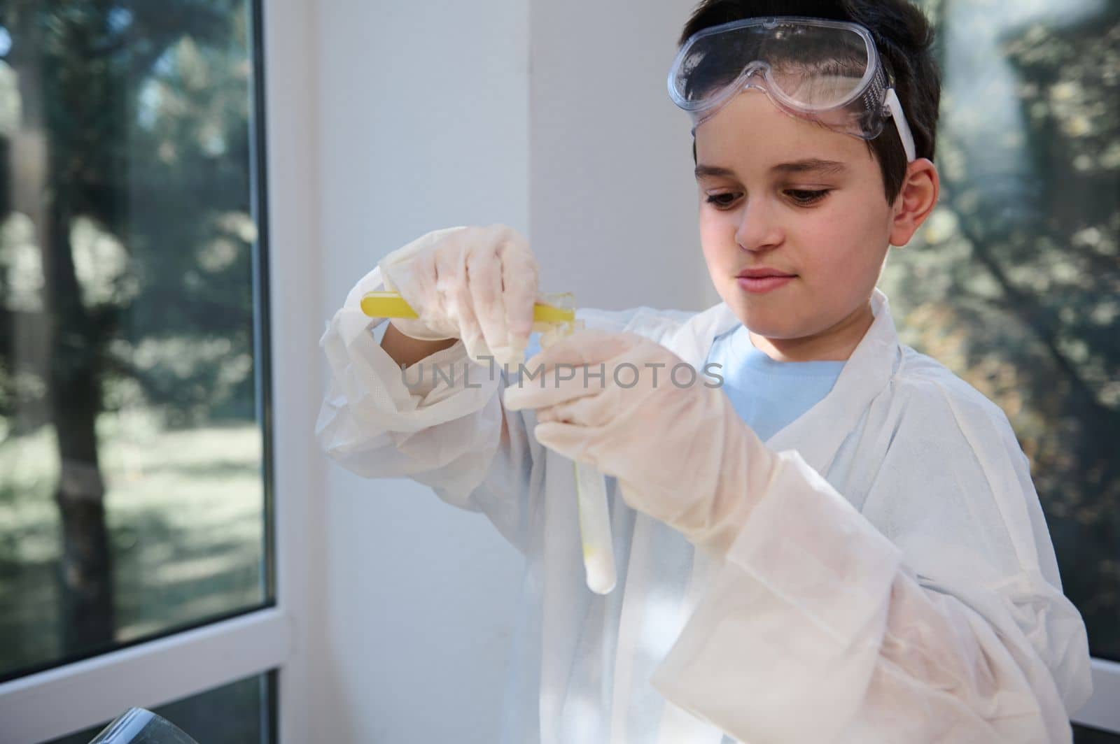 Smart teenage boy at chemistry class. Young chemist scientist, wearing white lab coat and protective gloves, pouring yellow liquid into a test tube, conducting chemical experiment in school laboratory