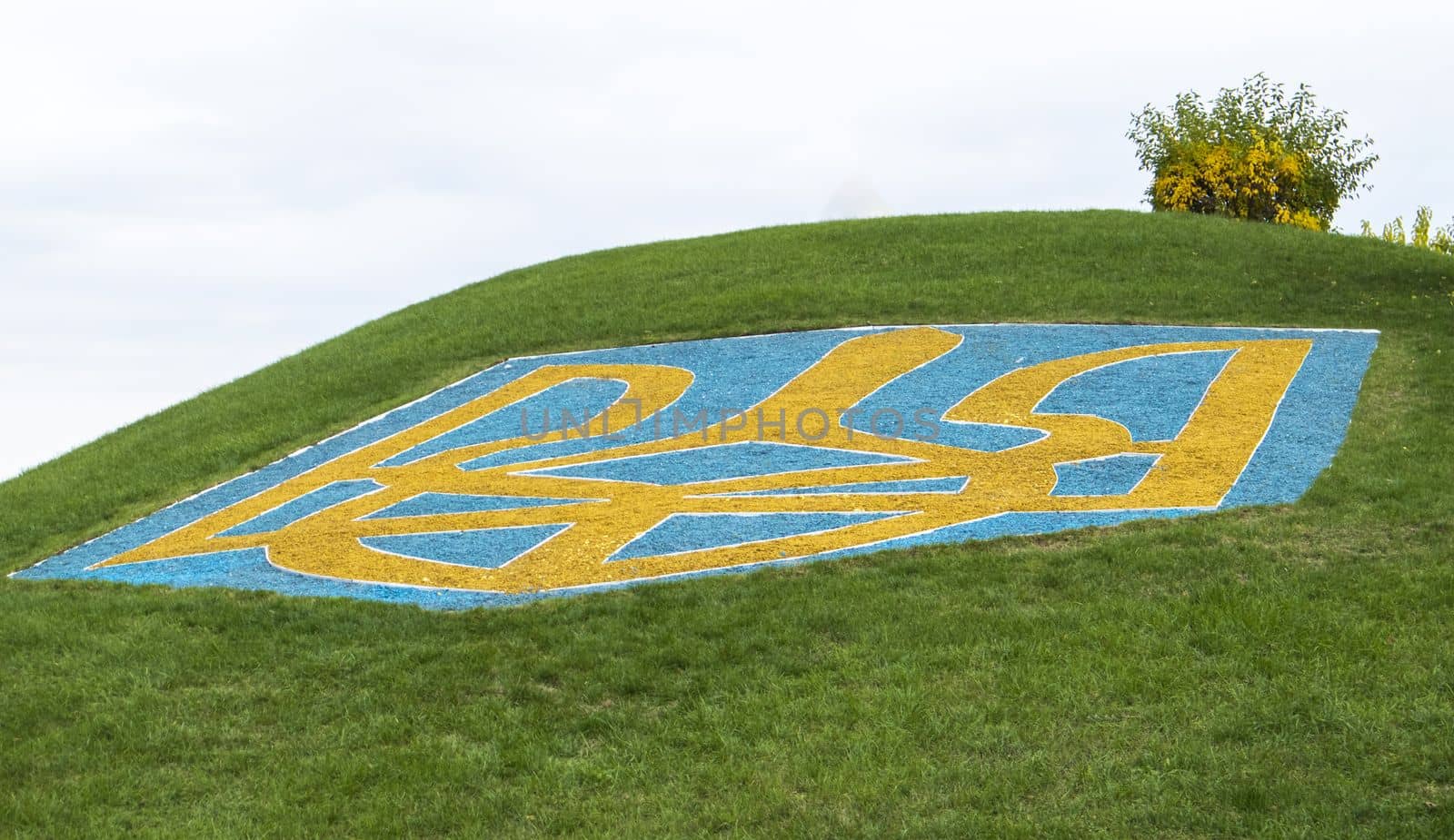 The large coat of arms of Ukraine on a green lawn lined with blue and yellow stones against a blue sky. The official symbol of the state. Trident of Prince Vladimir. Ukraine, Kyiv - October 08, 2022