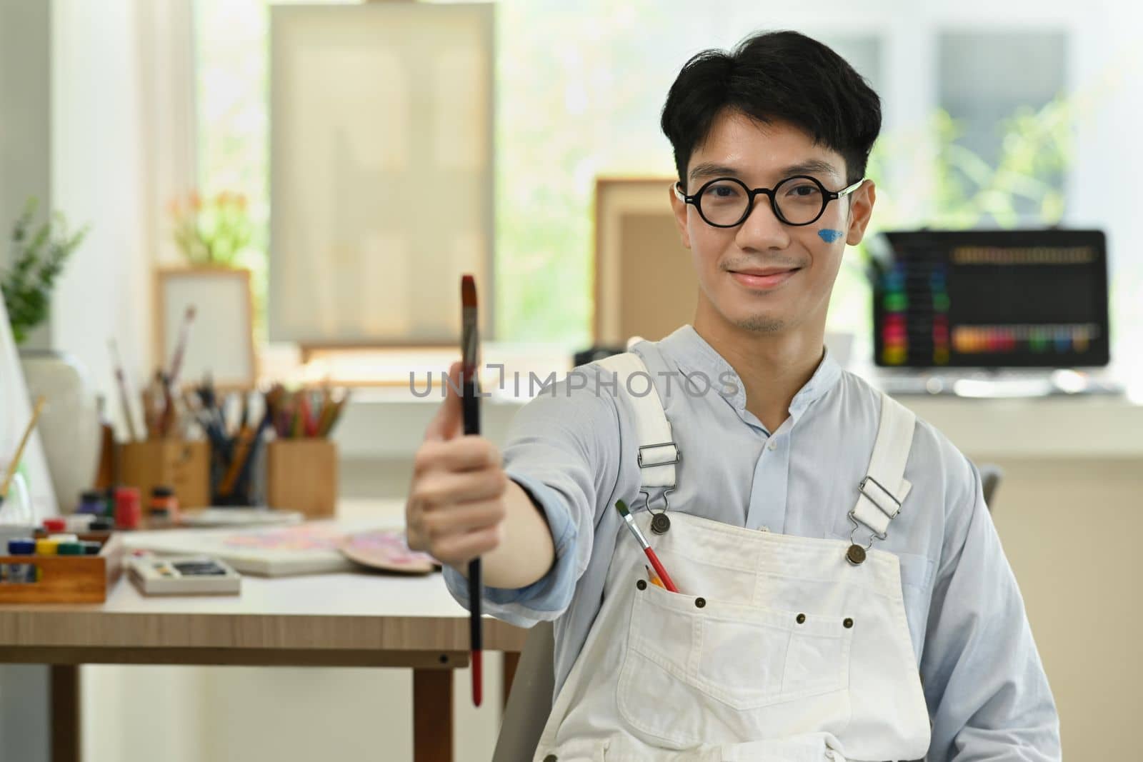 Handsome artist man holding paintbrushes and palette smiling to camera while sitting in art studio.