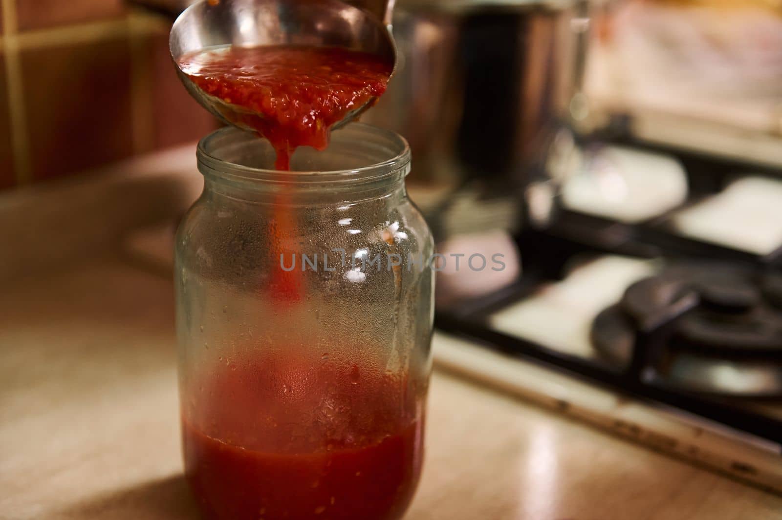 Close-up housewife in black chef's apron using a ladle, pours boiling tomato juice from saucepan into sterilized jar by artgf