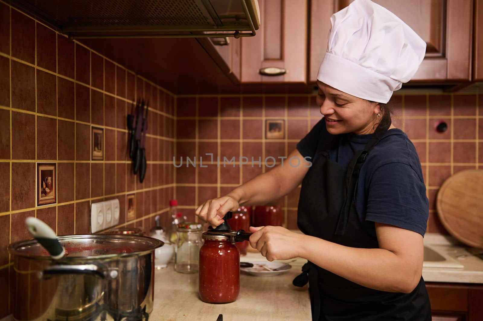 Pretty housewife in white chef's cap and apron, standing at a kitchen countertop, closing lids of jars with freshly brewed tomato juice, using a special seaming key, preserving food for winter