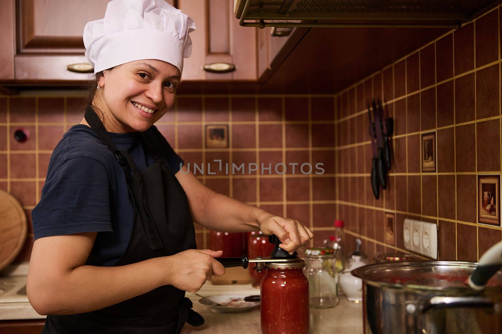 Smiling woman housewife in white chef hat and apron, using seamer, closes lids of jars with freshly canned tomato sauce, juice or passata, preserving food with organic homegrown tomatoes for winter