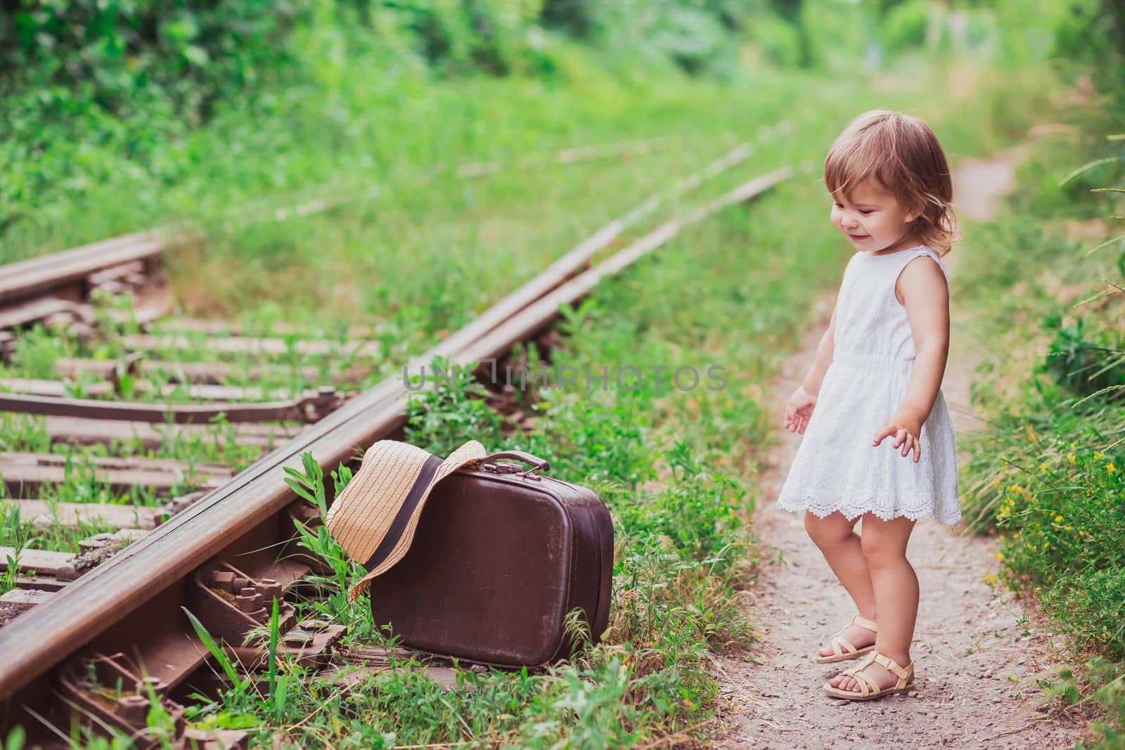 Charming baby with a suitcase walks along the railroad.