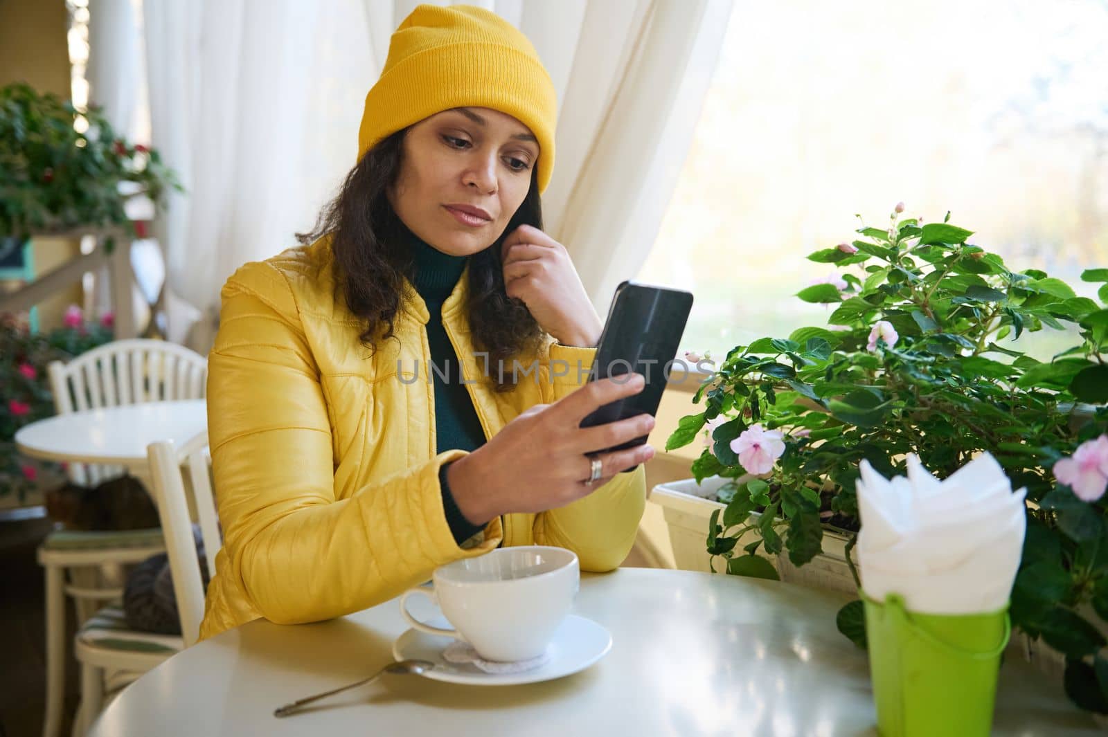 Charming gorgeous Latin American woman in a bright yellow coat and hat, sitting in a cafe enjoying a coffee break, and using her smartphone checking newsfeed, posting on social media. Autumn mood.
