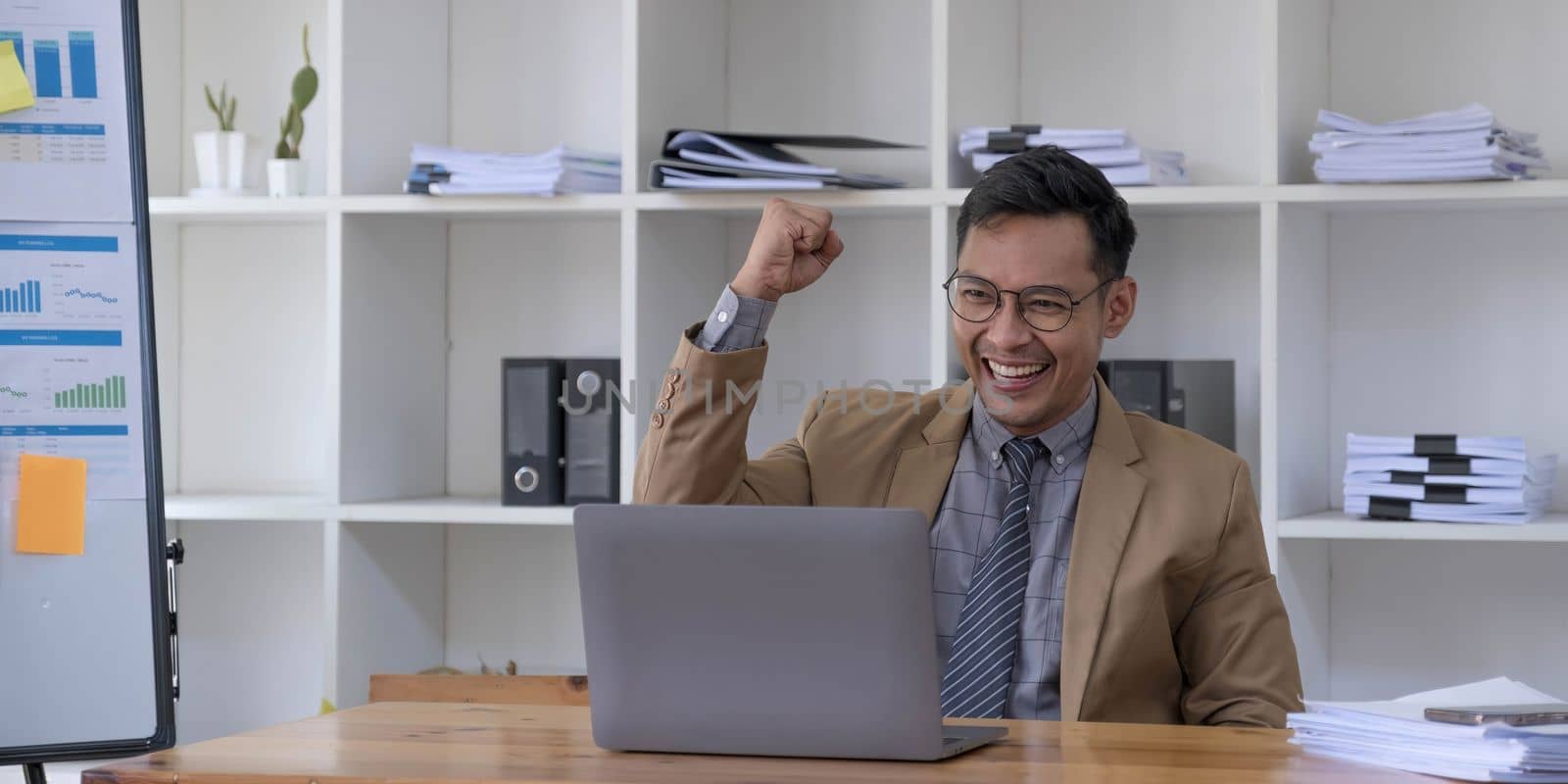 Happy young businessman in suit looking at laptop excited by good news online, lucky successful winner man standing at office desk raising hand in yes gesture celebrating business success win result by wichayada