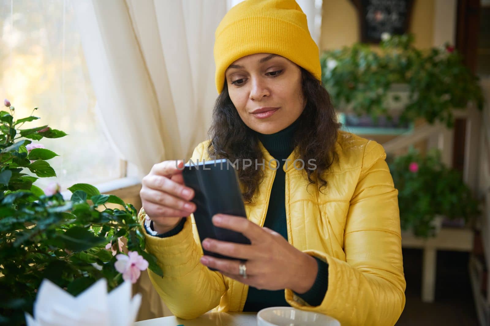 Close-up portrait of multi-ethnic pretty woman in bright yellow jacket and hat, using smartphone, checking news feed, scrolling web pages, checking social media content while chilling in a coffee shop