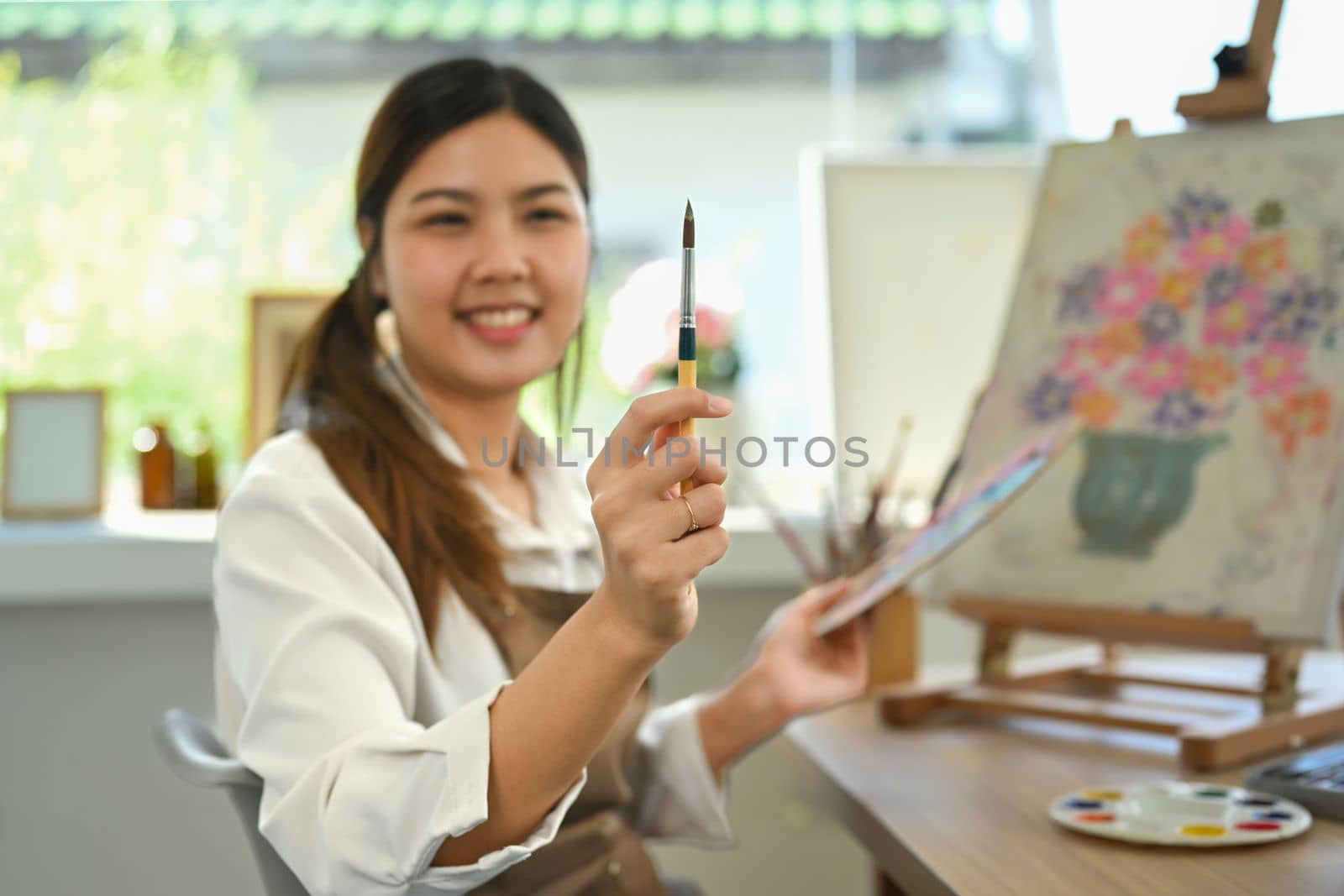 Pleasant female artist in apron with palette and brush painting on canvas at art studio. Education, hobby, art concept by prathanchorruangsak