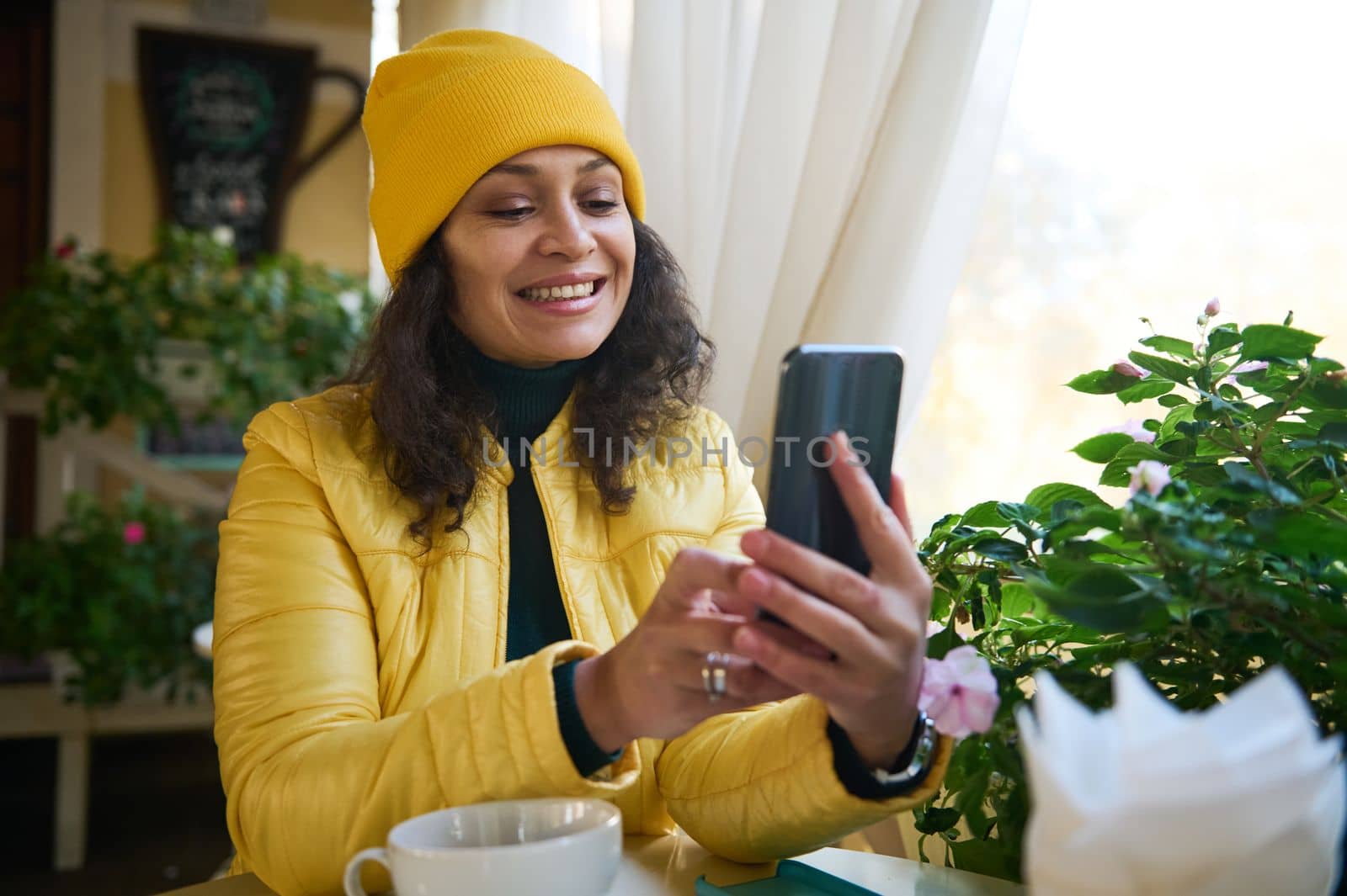 Charming dark-haired multi-ethnic woman in yellow clothes, smiling a beautiful toothy smile while having a video call on her smartphone, sitting by table in a cafe, enjoying her coffee break outdoors