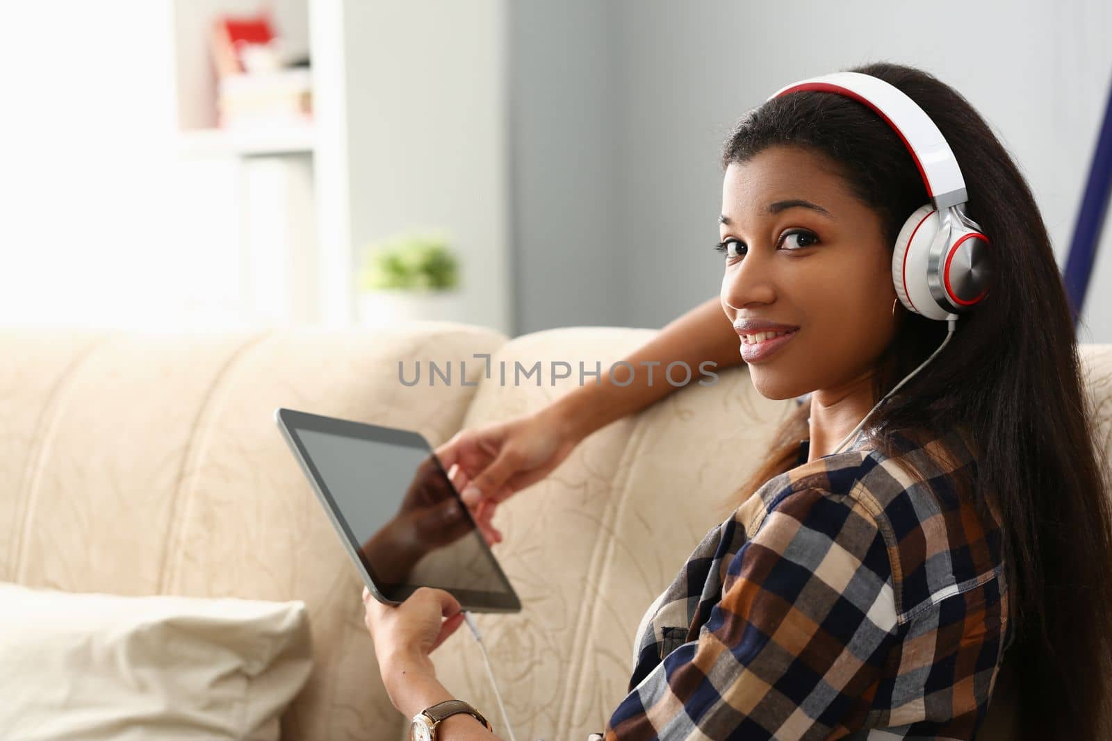 Young beautiful african american woman in headphones uses tablet. Listens to music looks at camera and smiles on sofa in room