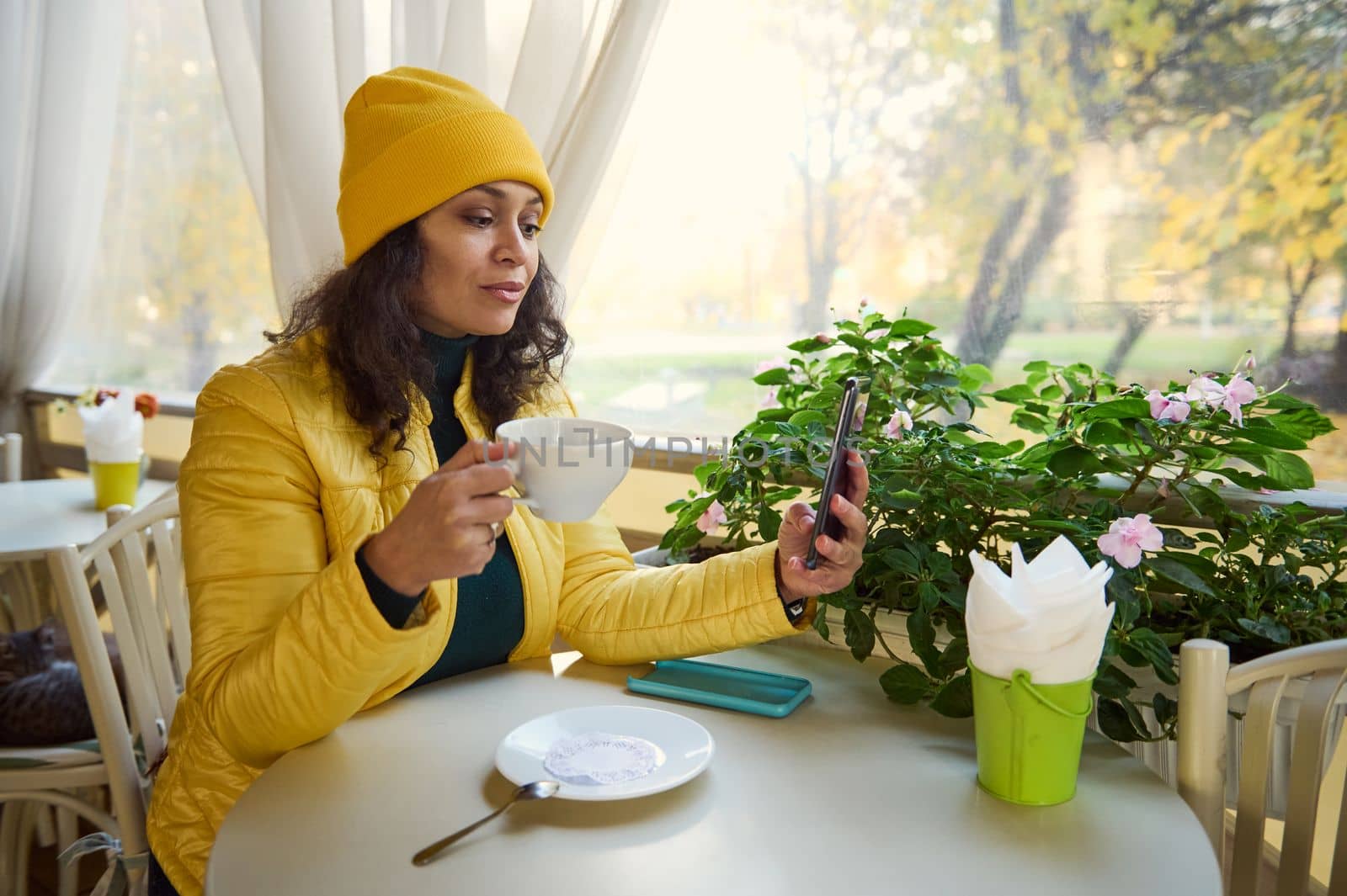 Charming Latin American woman in green sweater and yellow hat and jacket, checking news feed on her smartphone while resting in a coffee house, enjoying her hot espresso on a warm sunny autumn day