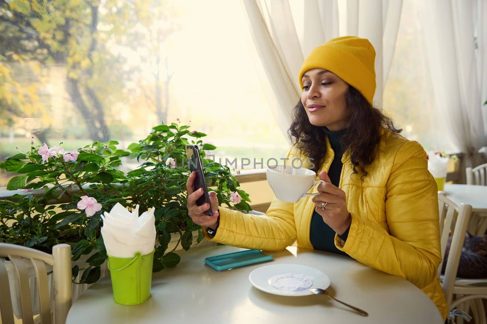 Happy stylish Latin American woman in bright yellow clothes, having coffee and reading news on mobile phone, checking her social media content in the autumn morning, enjoying her weekend outdoors