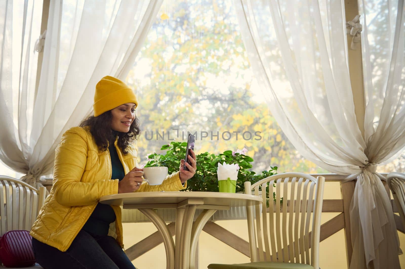 Beautiful smiling African American woman customer, dressed in bright yellow hat and jacket, sitting at table with hot coffee while talking on smartphone by video link in cozy cafe on sunny autumn day
