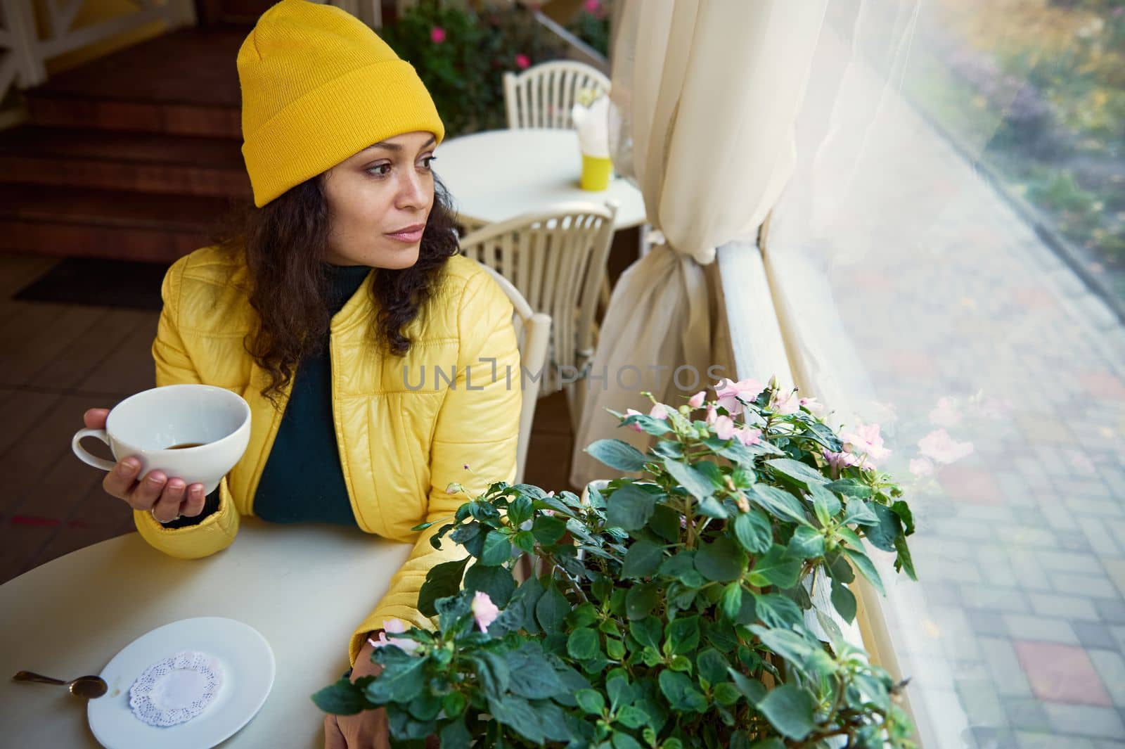 Portrait of a beautiful delighted woman in a bright yellow hat and jacket, holding a white ceramic cup of hot aroma coffee, looks out the window at the autumn park while chilling in a cozy coffee shop