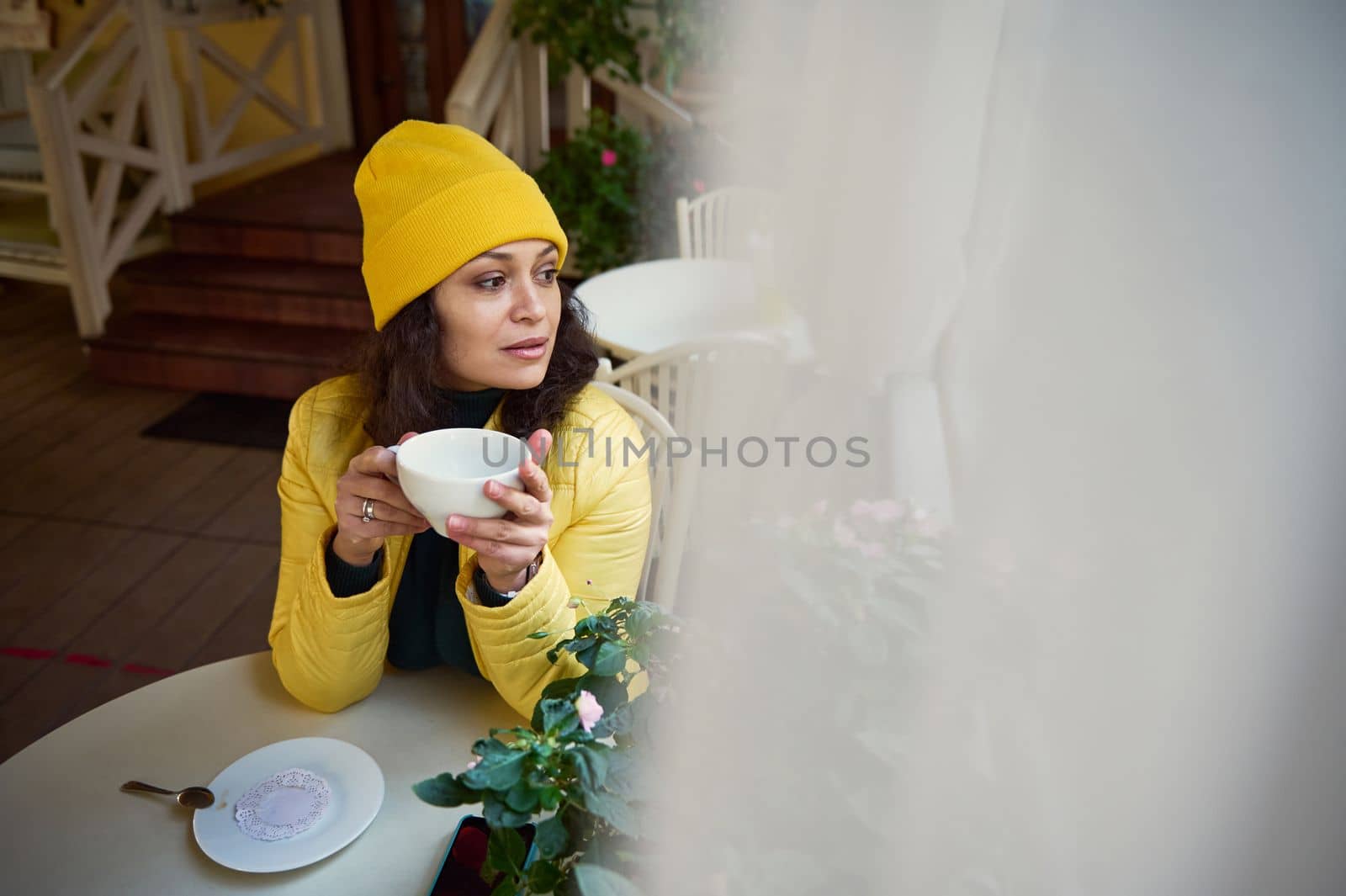 Attractive millennial curly brunette woman in a bright yellow hat and jacket, thoughtfully looking aside, relaxing in a cafe, taking a sip of hot aroma coffee. Blurred white curtains in the foreground
