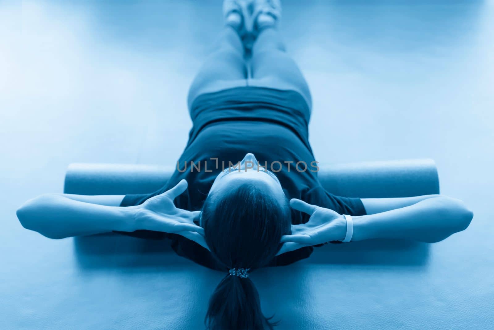 Attractive female doing foam roller exercise relaxing her back and posing in modern bright fitness center by Mariakray