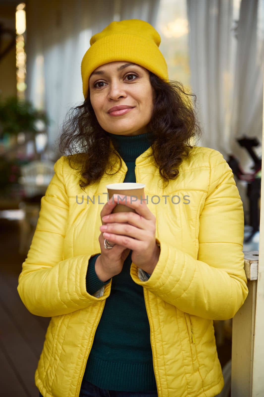 Confident portrait of a delighted attractive Middle-Eastern curly brunette woman, dressed in green sweater, yellow hat and jacket, warming her hands with a hot takeaway coffee in disposable paper mug