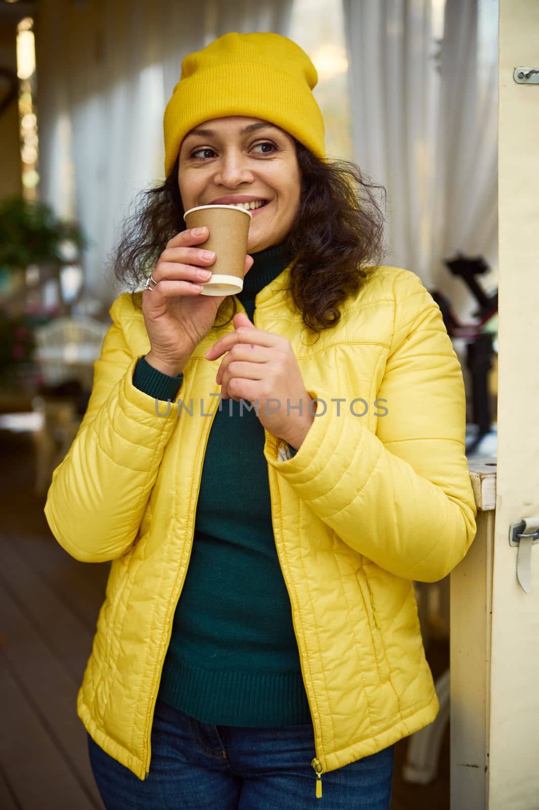 Waist-up portrait of multi-ethnic charming woman, dressed in green warm sweater, yellow wool hat and jacket, smiling, looking aside while enjoying her coffee break, taking a sip of hot aroma coffee