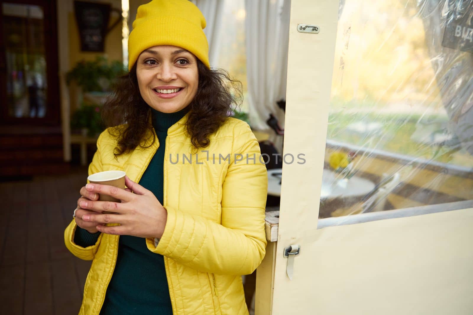 Charming positive African American young woman, in bright yellow hat and jacket, warming her hands with a takeaway hot coffee in a disposable paper cup, smiling a beautiful toothy smile looking aside