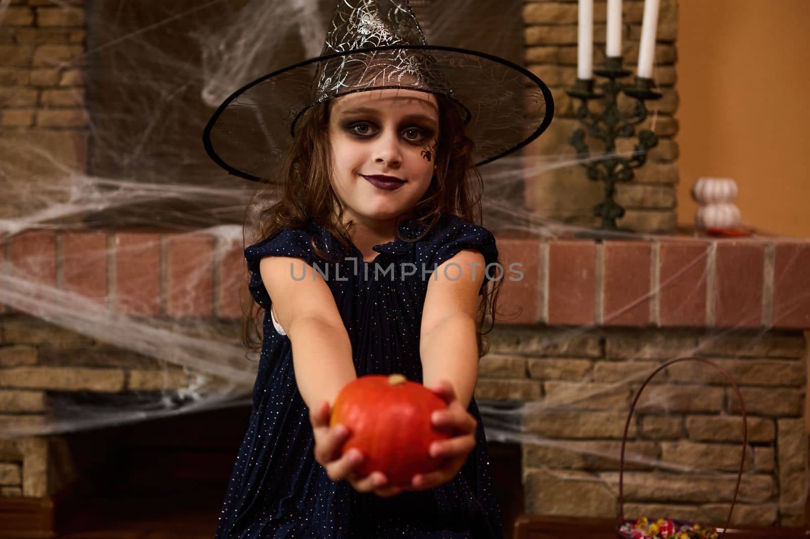 Adorable little girl in witch dress and wizard hat with art make-up on her face, handing a pumpkin to camera, sitting against a cobweb-covered fireplace, in a gothic home interior. Happy halloween