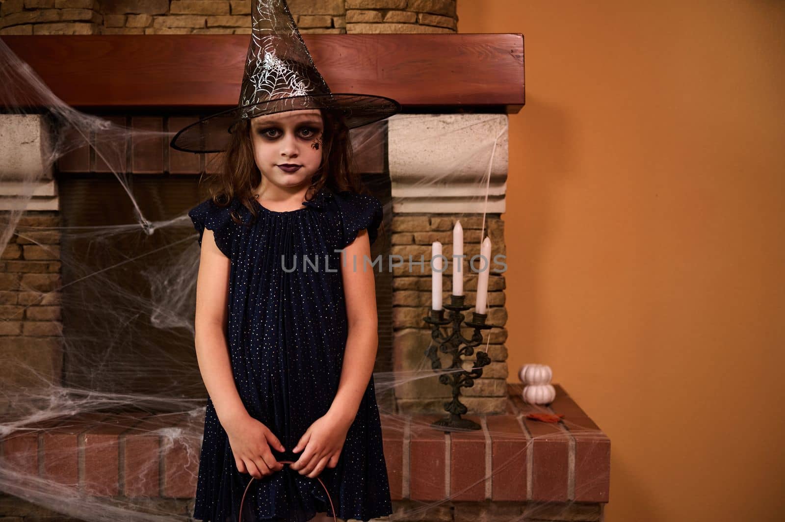 Mischievous mysterious Caucasian little child girl in witch costume and wizard hat, stands against a cobweb covered fireplace and candles, ready for Halloween party. Halloween trick or treat