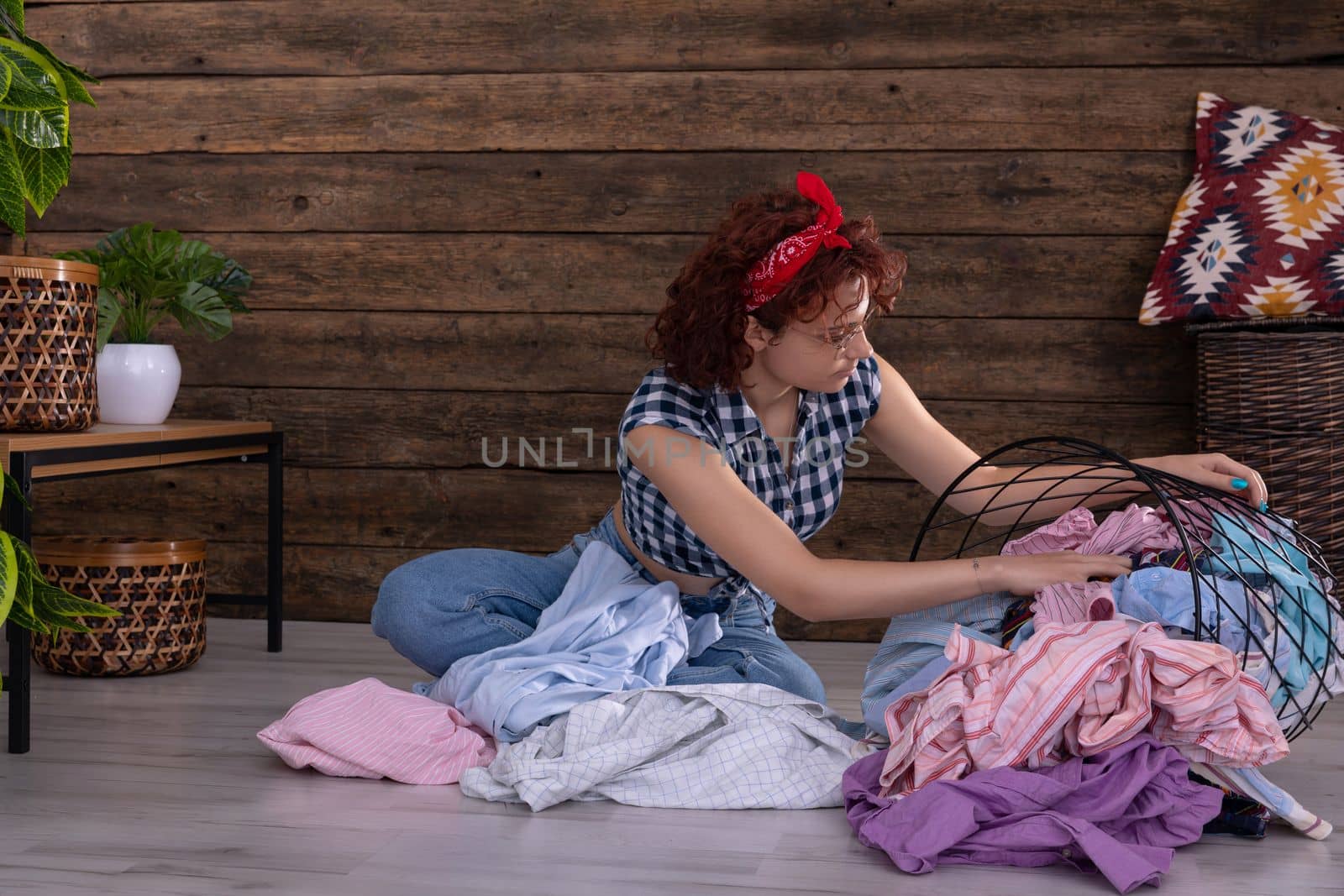 Attractive girl sorts clothes for washing. Short curly hair. Metal basket full of clothes prepared for washing. A wall of brown boards.