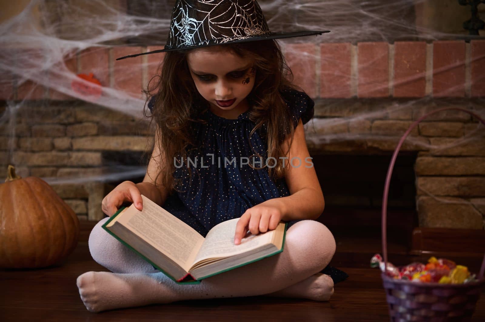 Little girl enchantress in wizard's hat, surrounded by Halloween treats casting spells while reading a sorcery book. by artgf