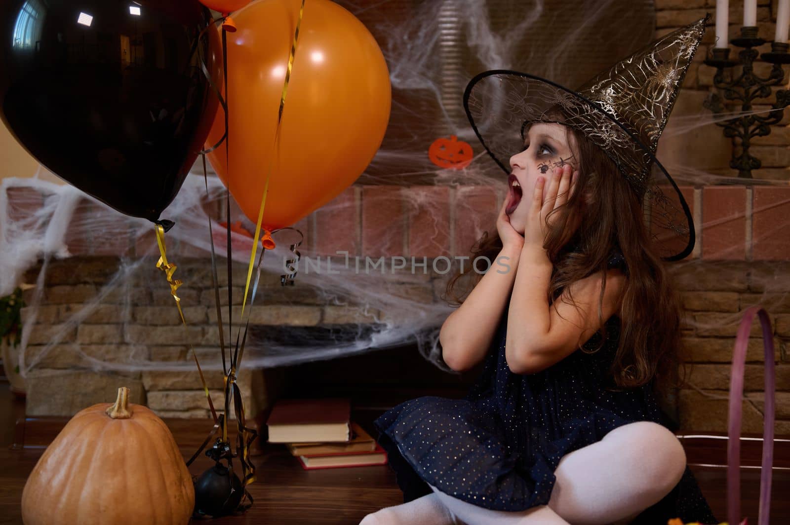 Surprised Caucasian little girl, dressed in witch and wizard hat, looking at orange black balloons while sitting against a cobweb-covered fireplace. Halloween party. Gothic festivity. October 31