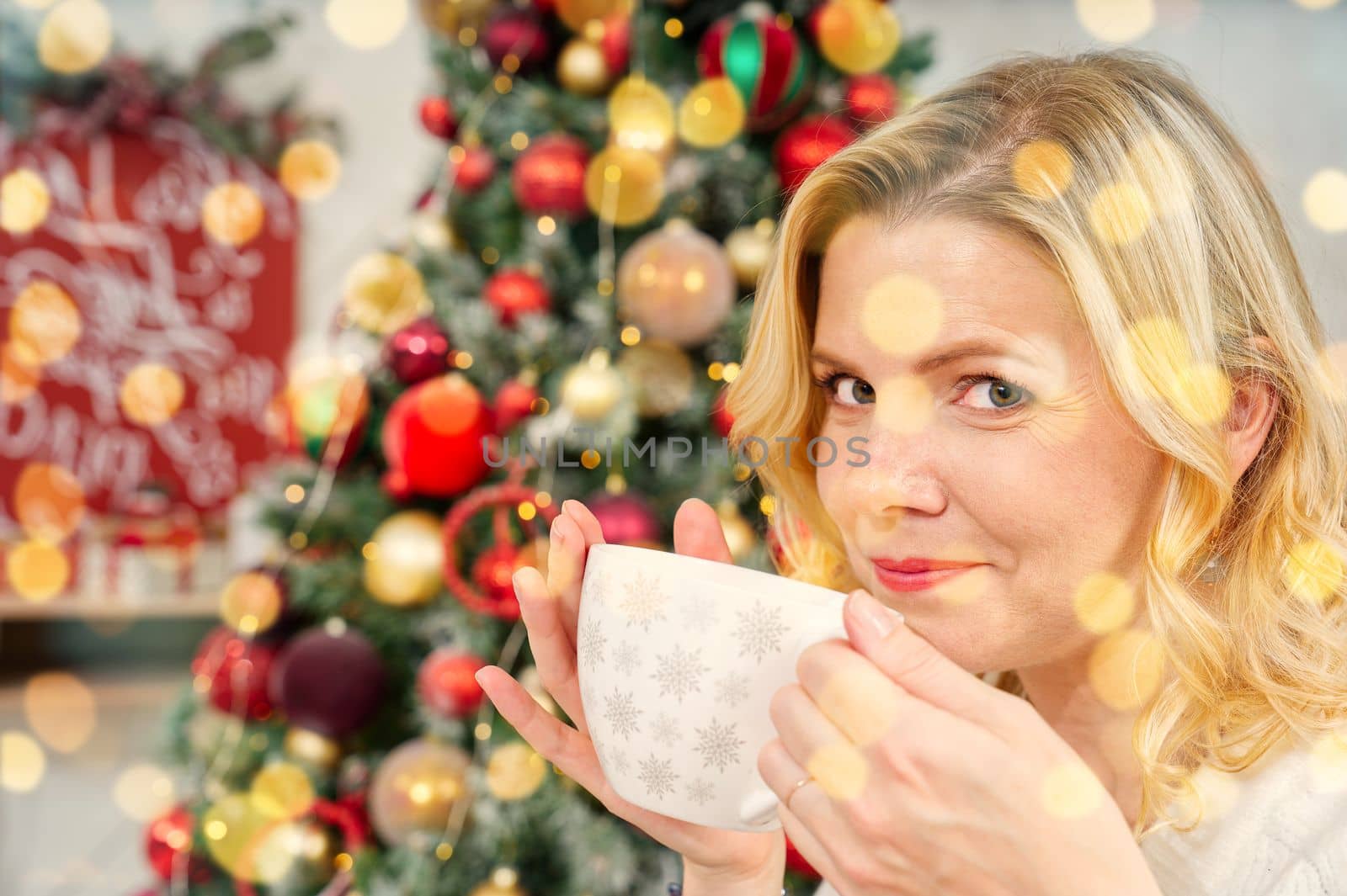 Girl in white sweater holding a cup of warm coffee or tea on background of a Christmas tree. by PhotoTime