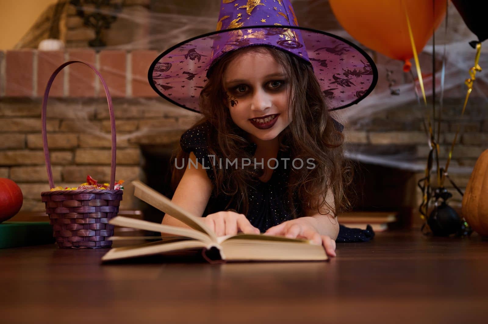 Mysterious little girl witch in wizard's hat, looking at camera while learning sorcery, reading a spell book, lying on the floor against a spider web covered fireplace, surrounded by Halloween treats