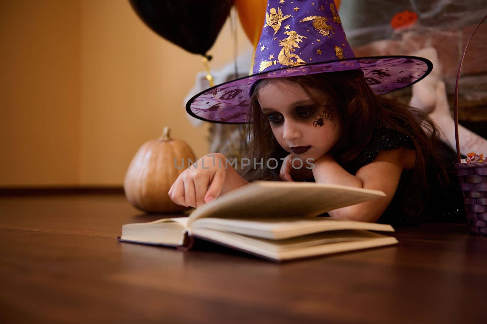 Mischievous little girl with Halloween make-up dressed as a witch, a sorceress in a wizard's hat reading a book of witchcraft and magic spells surrounded by pumpkins and a festive gothic atmosphere